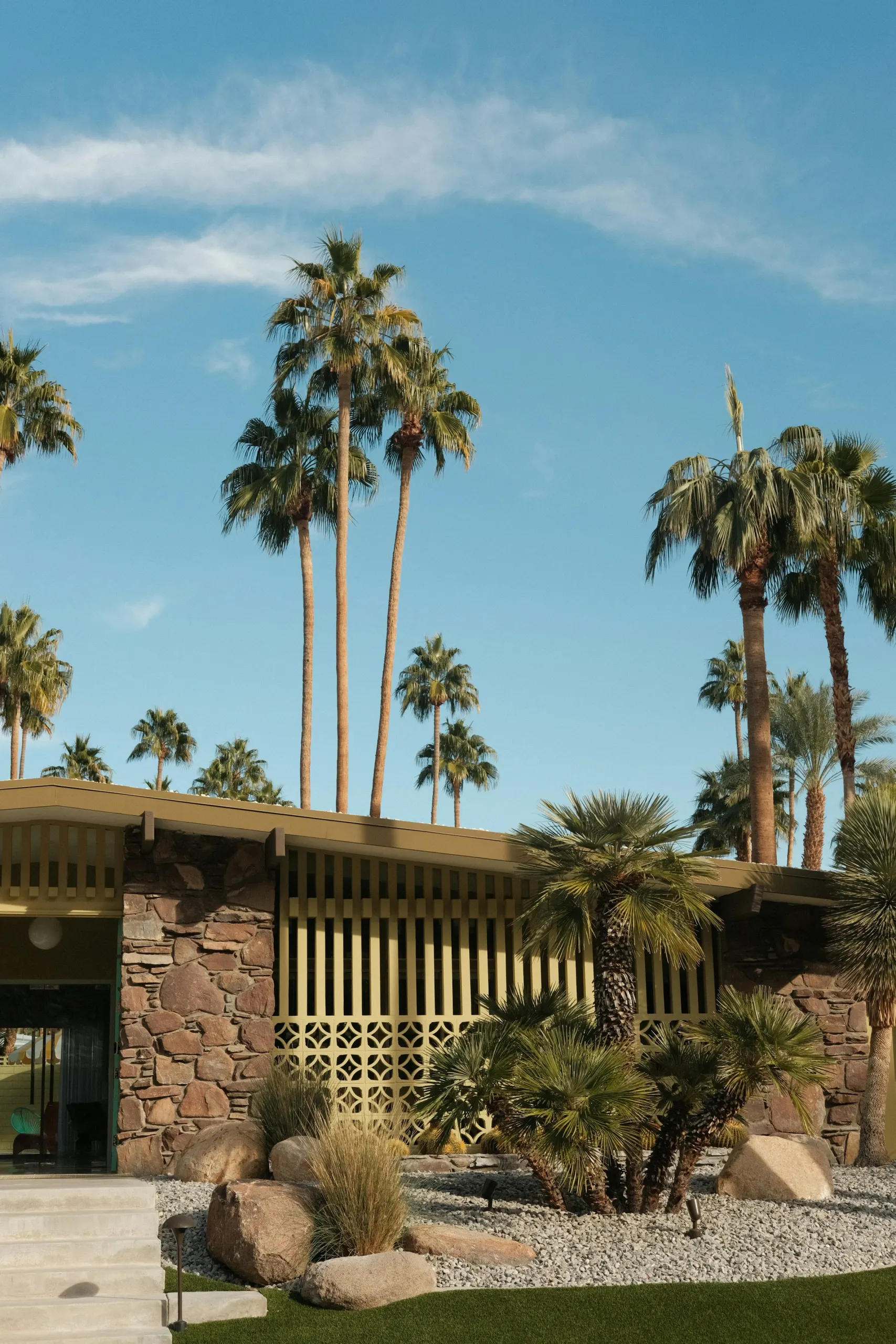Mid-century home with stone walls and tall fan palms against a clear sky