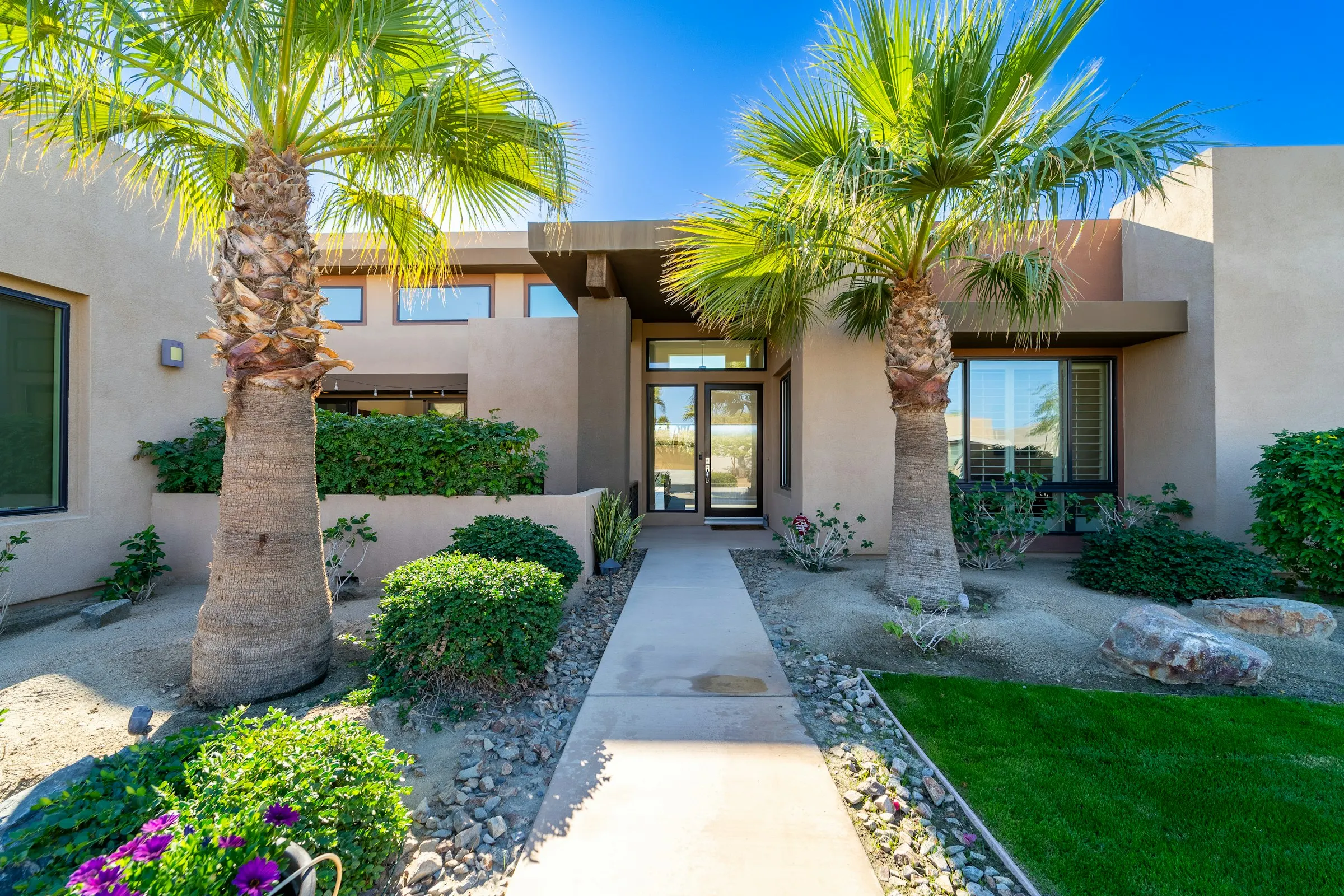 Front walkway lined with palms, rounded shrubs, and flowering beds leading to a stucco home